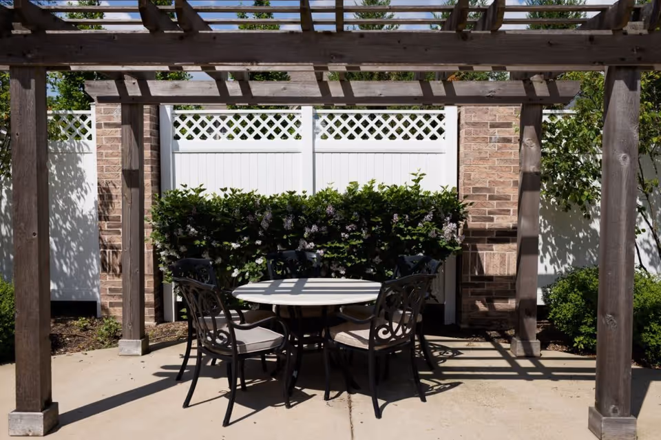 Outdoor patio under a wooden pergola with a round table and four chairs in front of a white fence and brick wall.