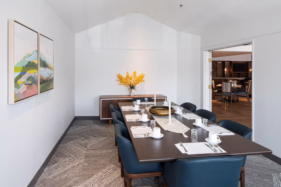 A modern dining room with a long dark wooden table set for eight people. The table has white placemats, cups, glasses, and cutlery. A decorative centerpiece with a brass bowl and two white candles is on the table. There is a sideboard against the back wall with a vase of yellow flowers. Two abstract paintings hang on the left wall. An open doorway leads to another room with additional dining tables and chairs.
