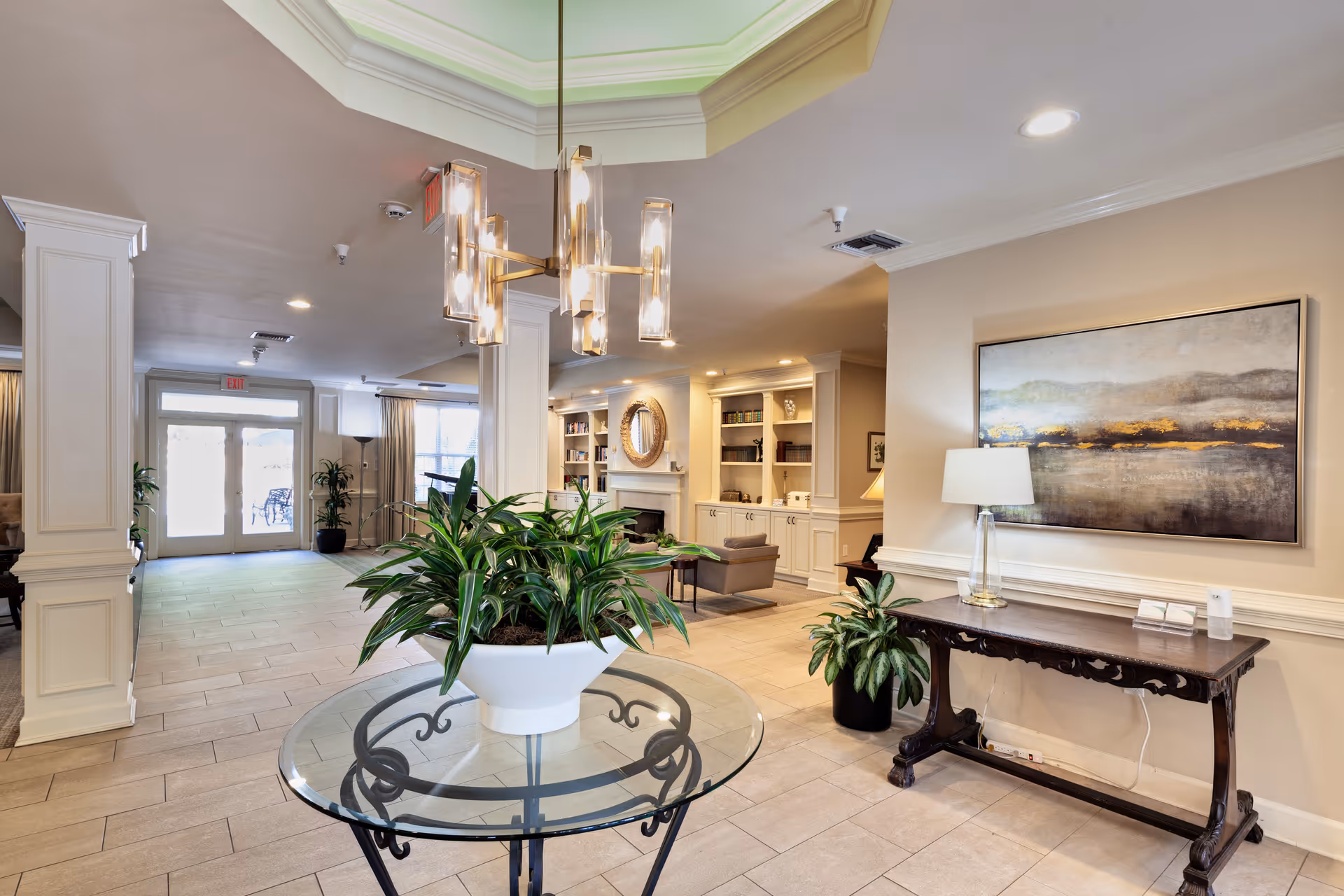 A bright and spacious senior living facility common area with a glass table holding a large potted plant in the center. The room features beige tiled flooring, cream-colored walls, a modern chandelier, and a decorative wooden console table with a lamp and artwork above it. In the background, there is a seating area with armchairs, built-in shelves, and a fireplace, as well as large glass doors leading outside.