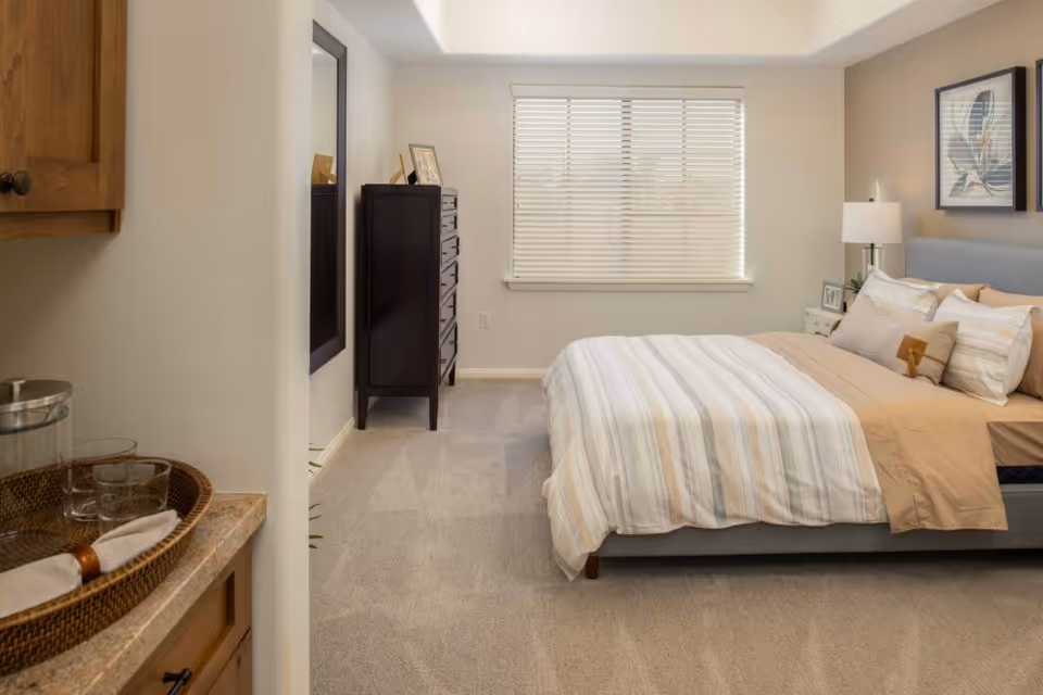 Sunlit furnished bedroom with a bed, nightstand, dresser and a window with blinds.