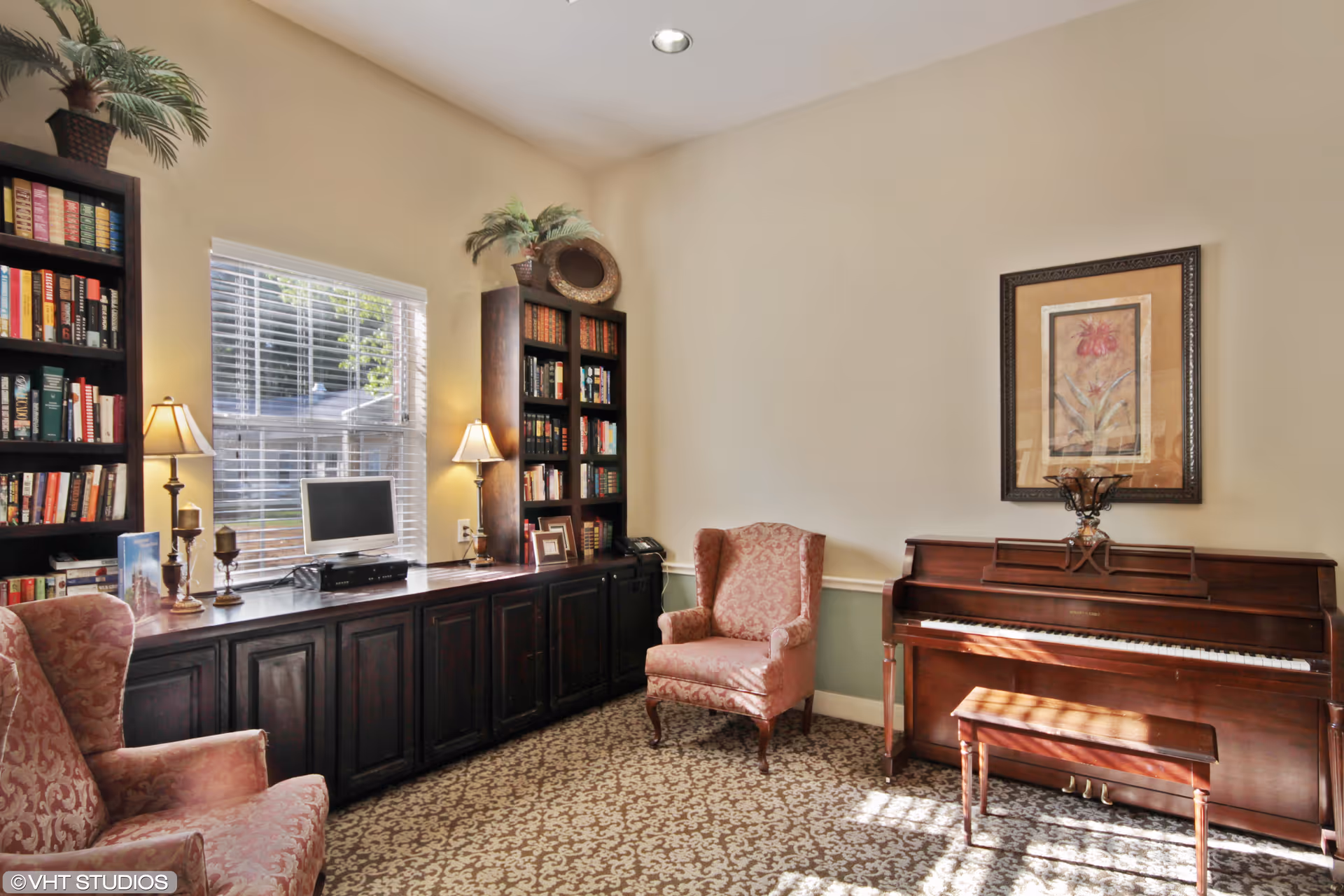 A cozy interior room with two upholstered armchairs, a wooden piano with a matching bench, two tall bookshelves filled with books, a window with blinds, and a long wooden cabinet with two table lamps and decorative items on top. A framed floral artwork hangs above the piano.