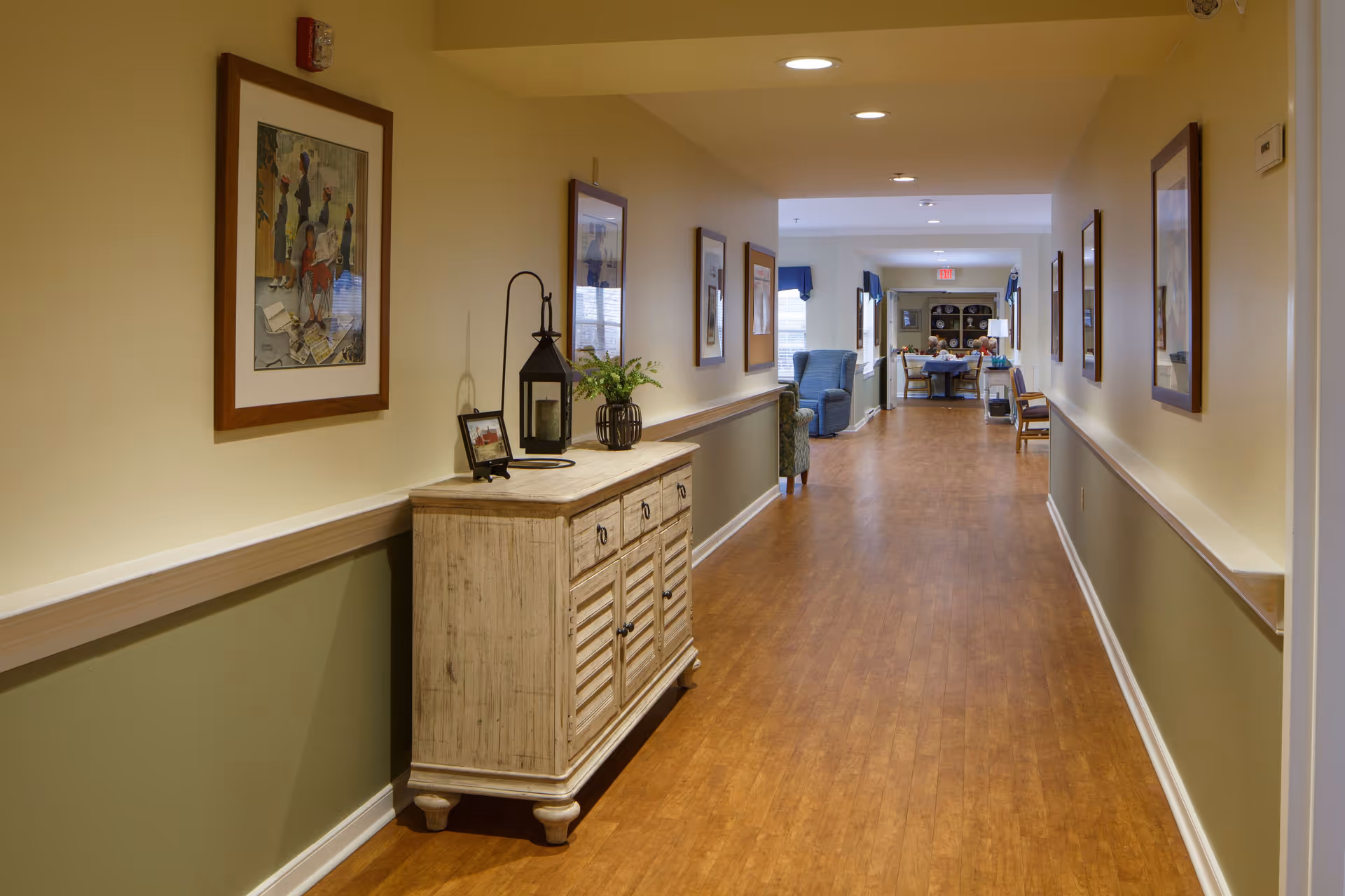 A well-lit hallway in a senior living facility with wooden flooring and beige walls. The hallway features framed artwork on both sides and a light-colored wooden cabinet with decorative items including a lantern and a small plant. At the end of the hallway, there are chairs and a table with more decorations near a window.