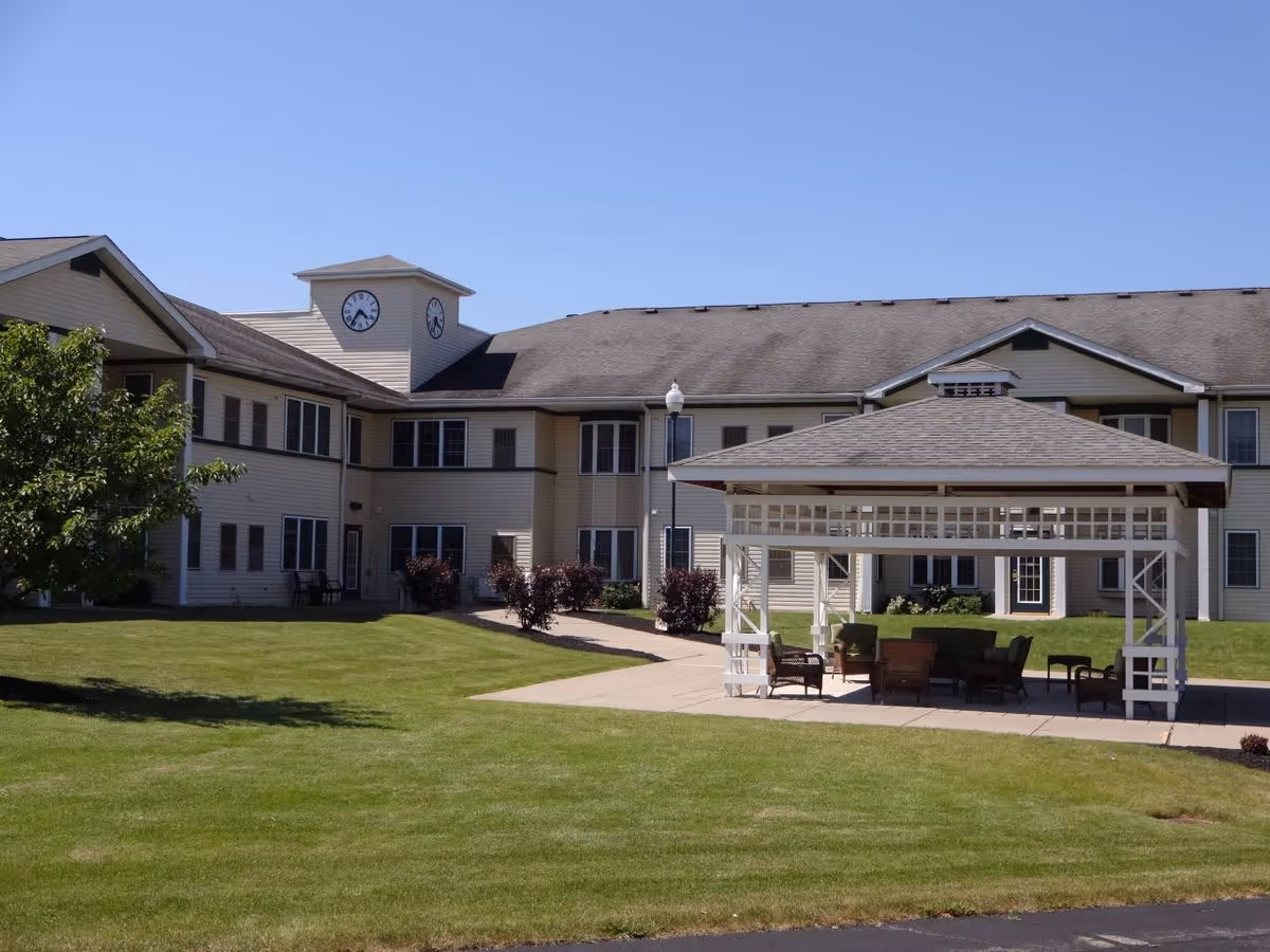 Two-story senior living building with a clock tower, green lawn, and a covered outdoor seating gazebo.