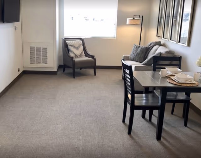 Small furnished living area with a sofa, armchair, dining table and chairs near a window and floor lamp.