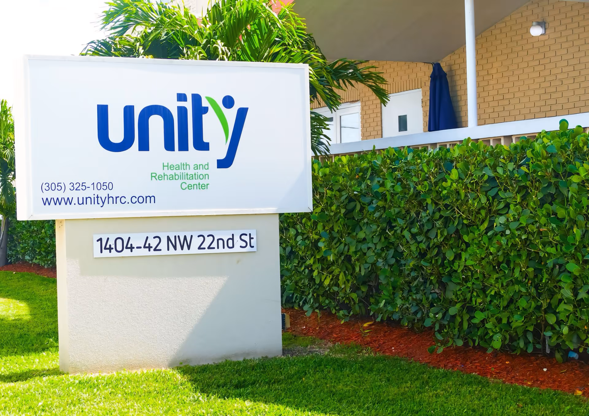 Outdoor sign for Unity Health and Rehabilitation Center showing the logo, phone, website and address in front of hedges and the building.