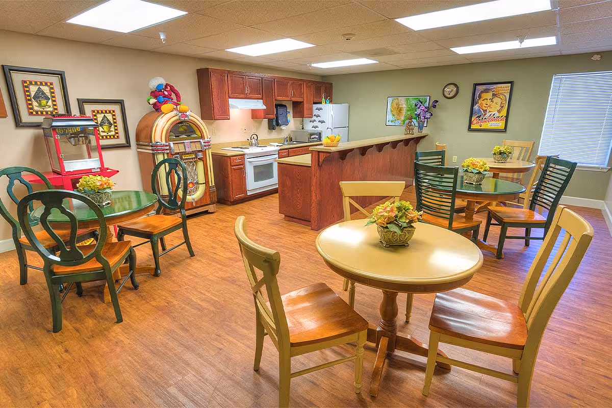 A cozy dining area with multiple round tables and wooden chairs, a kitchen with wooden cabinets, a white stove, and a refrigerator. The room features a vintage jukebox, a popcorn machine, framed artwork on the walls, and a window with blinds letting in natural light.