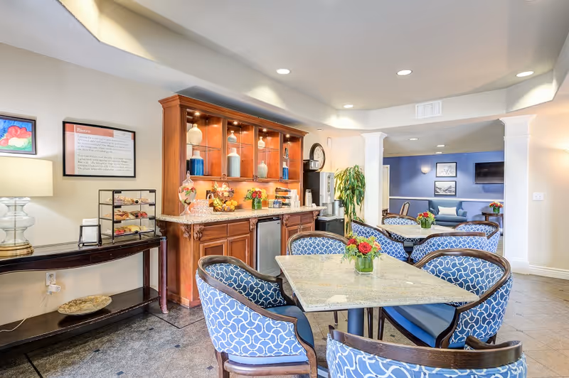 Bright common dining area with marble-top tables and patterned chairs in front of a wooden beverage and refreshment counter.