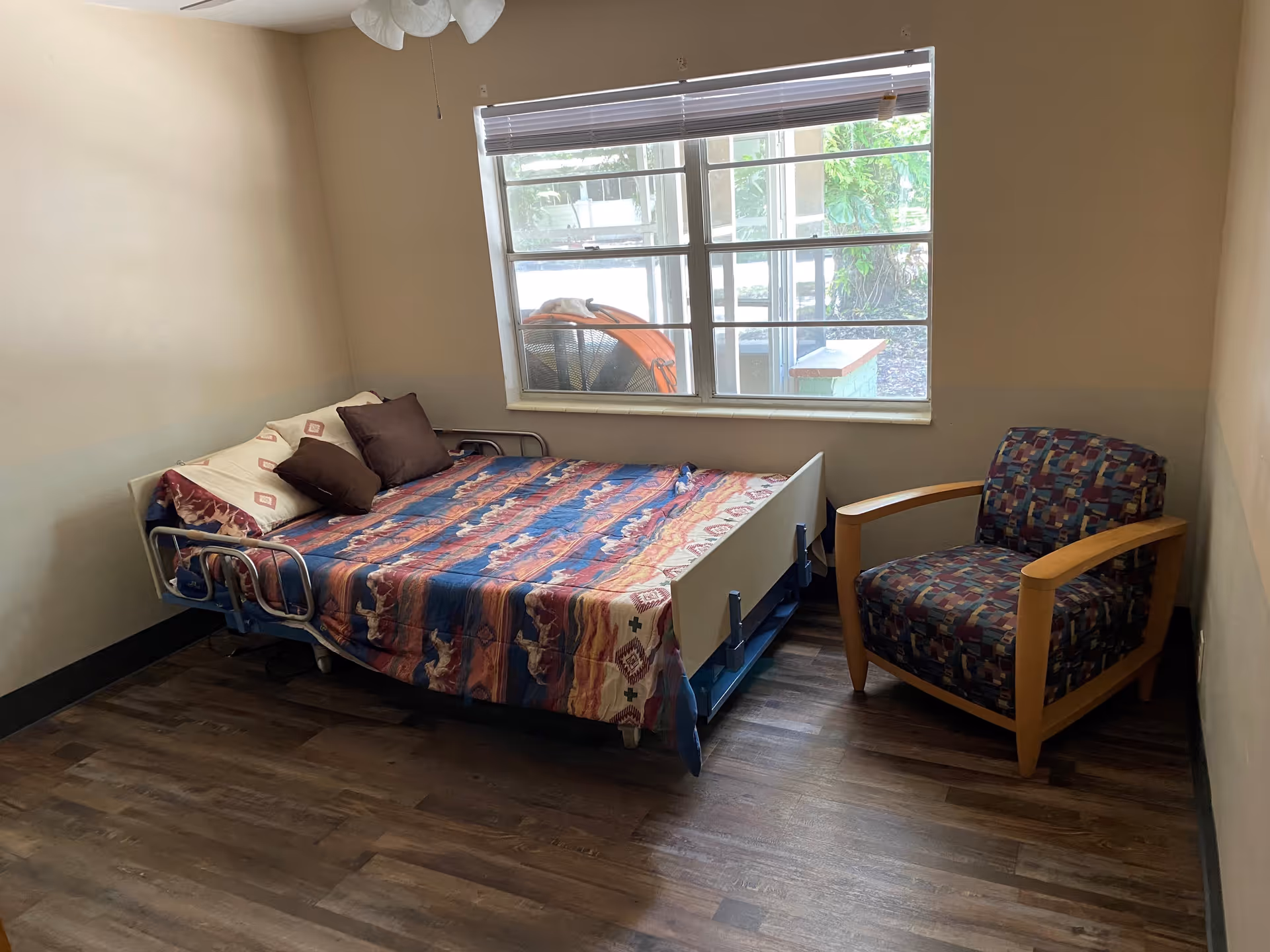 A small bedroom with a hospital-style bed covered with a colorful blanket and several pillows. Next to the bed is a cushioned armchair with wooden armrests. A large window with blinds lets in natural light, showing some greenery outside. The room has wooden flooring and beige walls.