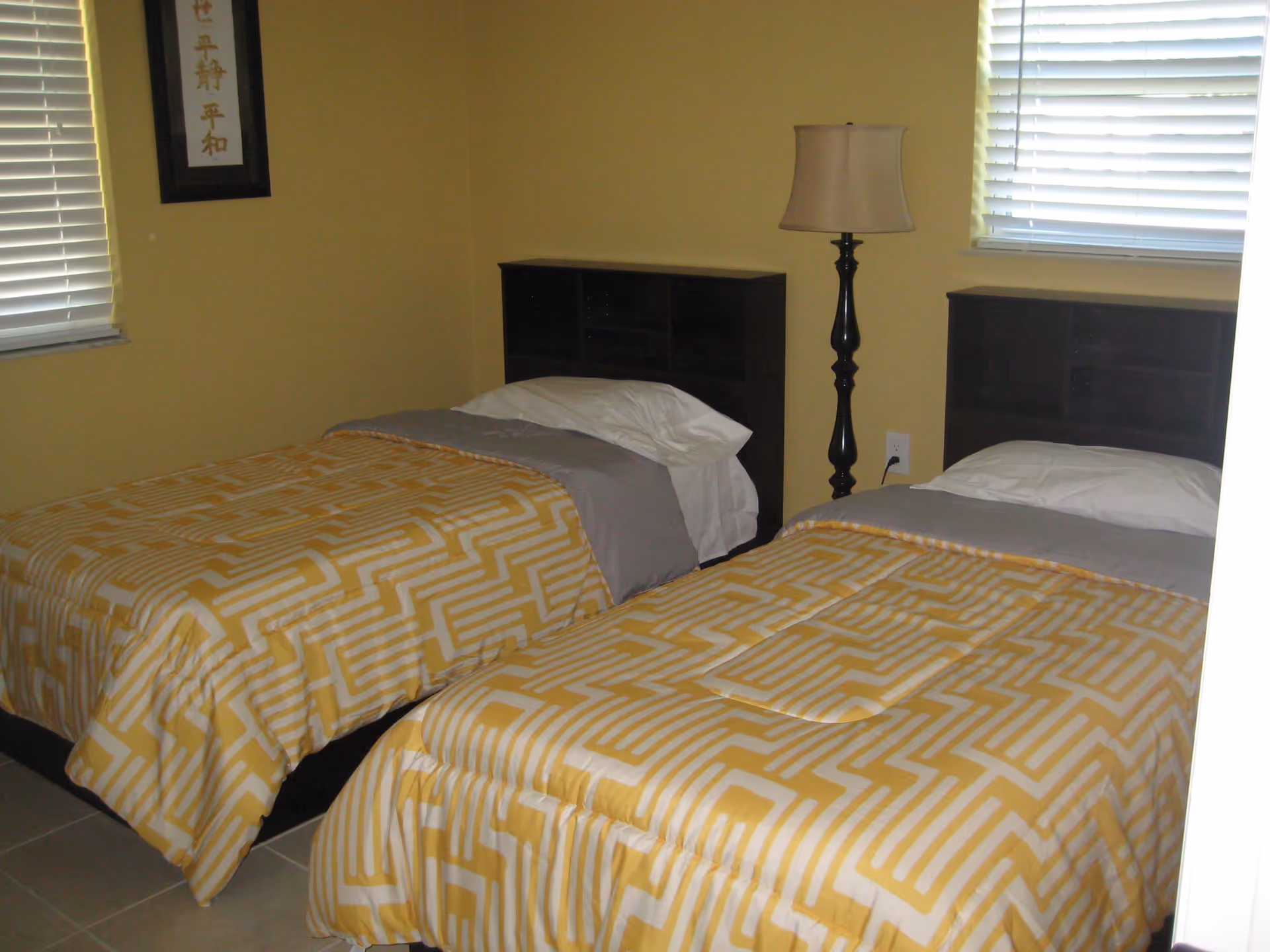 A bedroom with two twin beds featuring yellow and white patterned bedspreads and gray blankets. Each bed has a dark wooden headboard with shelving. There is a tall black floor lamp between the beds, and two windows with white blinds on the yellow-painted walls. A framed piece of art with Asian characters hangs on the left wall.
