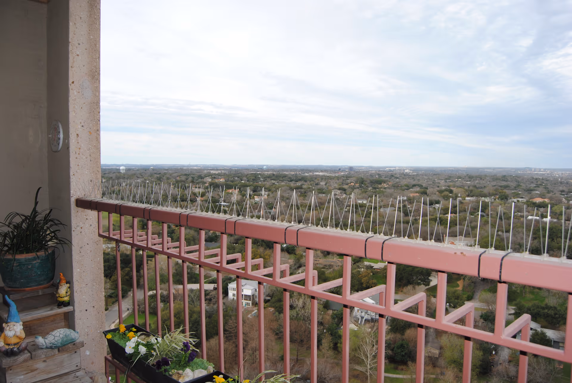View from a balcony with a pink railing overlooking a vast landscape of trees and houses under a cloudy sky. The balcony has potted plants and garden gnome decorations on a wooden shelf.