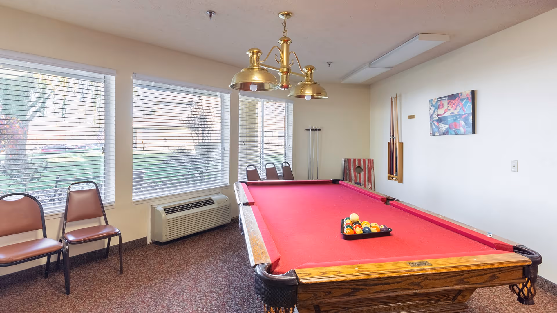 A recreational room with a red felt pool table in the center, a set of billiard balls arranged on the table, three brown chairs lined up against the wall with large windows showing an outdoor view, a wall-mounted rack holding pool cues, a cornhole board leaning against the wall, and a brass overhead light fixture above the pool table.
