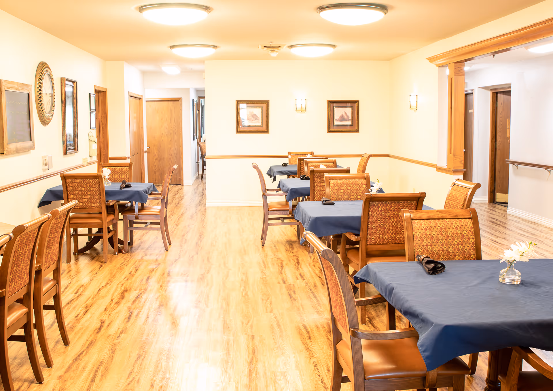 A bright dining room with wooden floors and several tables covered with blue tablecloths. Each table is surrounded by wooden chairs with patterned upholstery. The walls are light-colored with framed artwork and wall sconces. The room has ceiling lights and a hallway visible in the background.