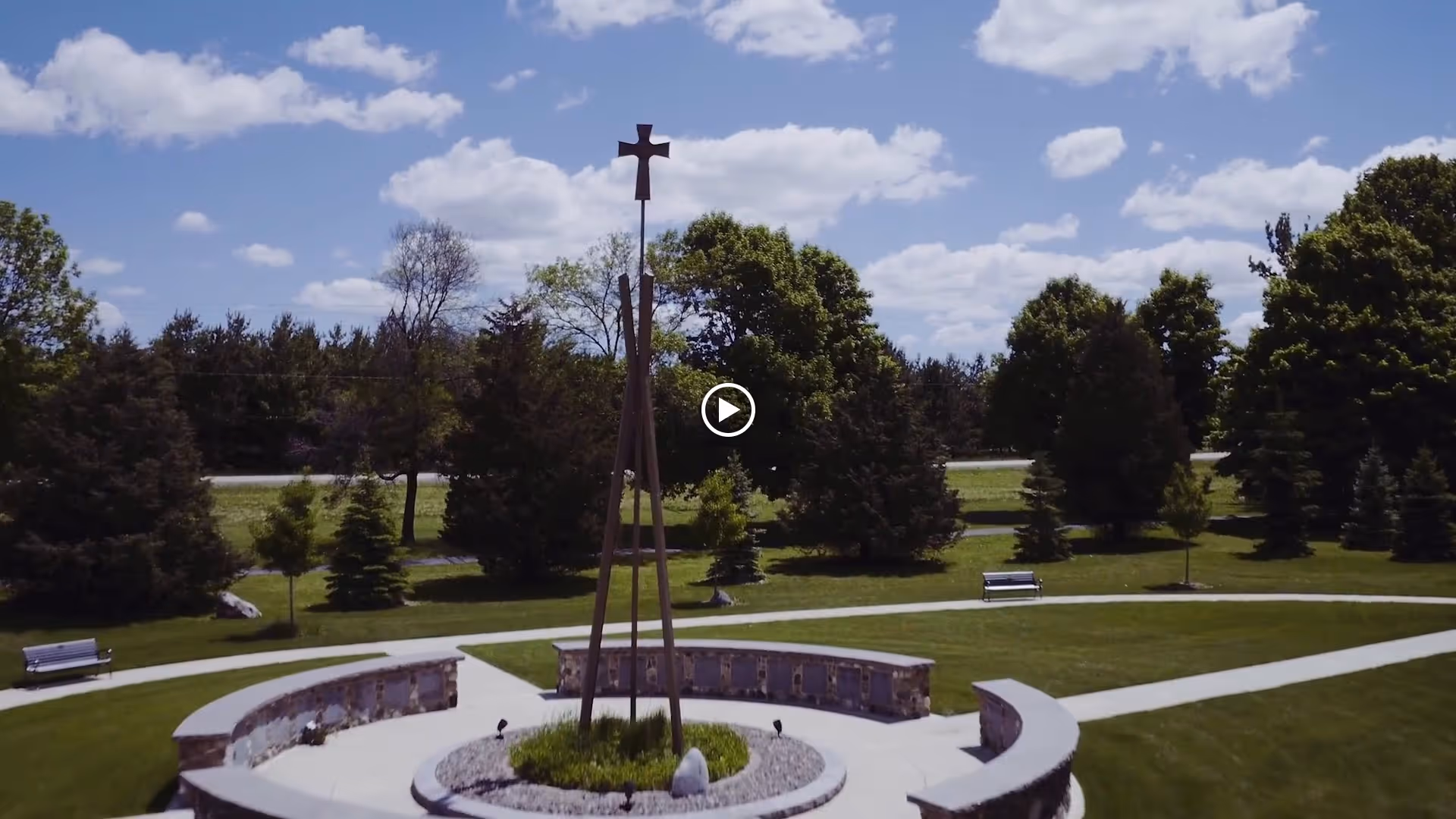 Outdoor circular memorial or garden area with a tall wooden cross structure in the center, surrounded by a curved stone bench and green grass. Trees and a partly cloudy sky are visible in the background.