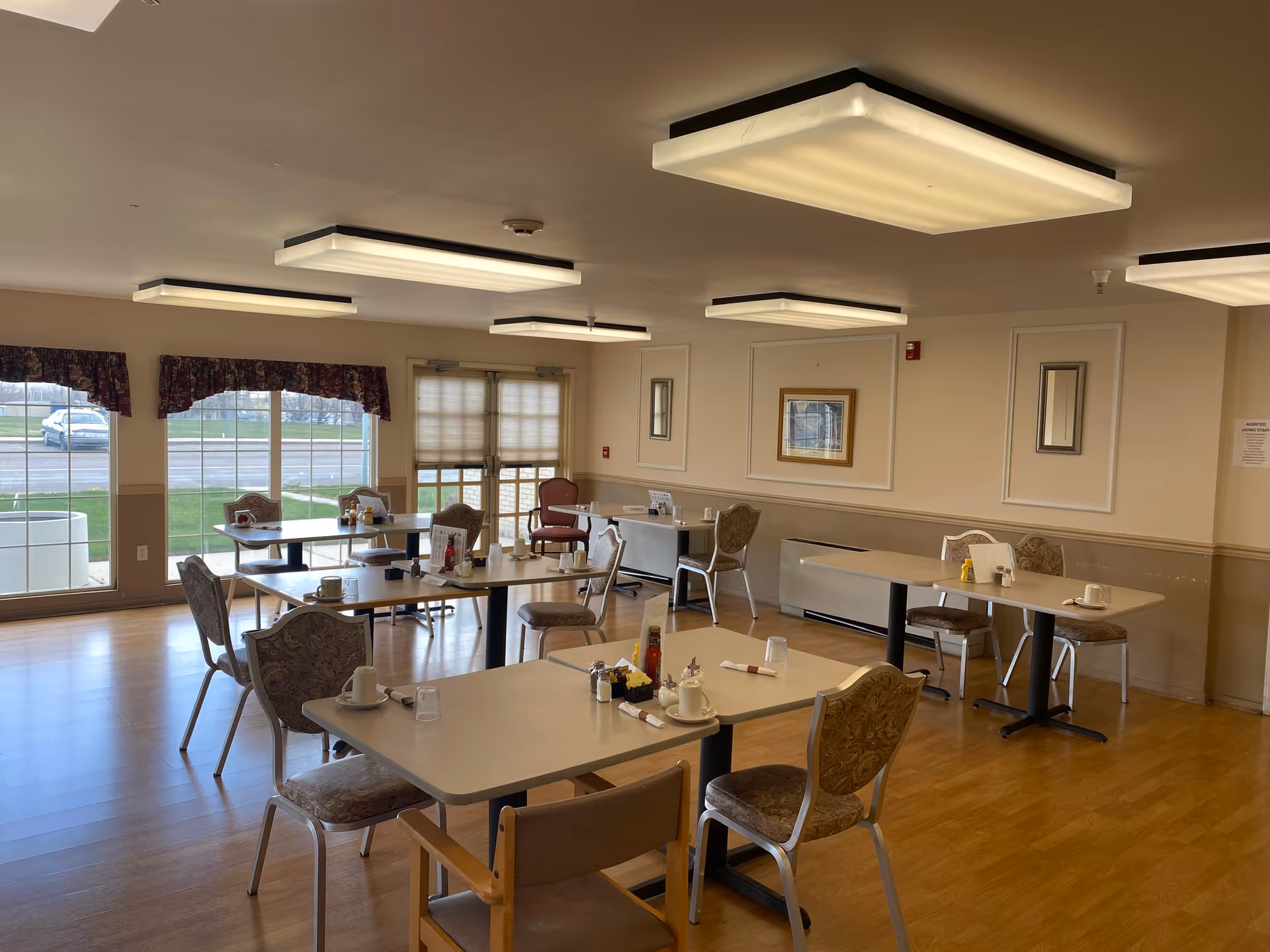 A dining room in Golden Eagle Plaza with several tables and chairs arranged neatly. Each table has cups, condiments, and napkins. Large windows and glass doors let in natural light, and the room has wood flooring and beige walls with framed artwork.