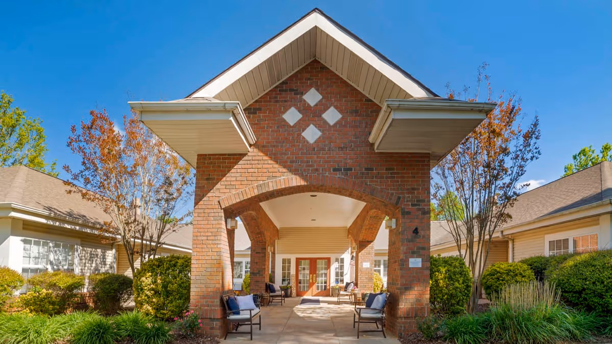 Entrance of a senior living facility with a brick archway and covered walkway, surrounded by well-maintained bushes and trees under a clear blue sky.