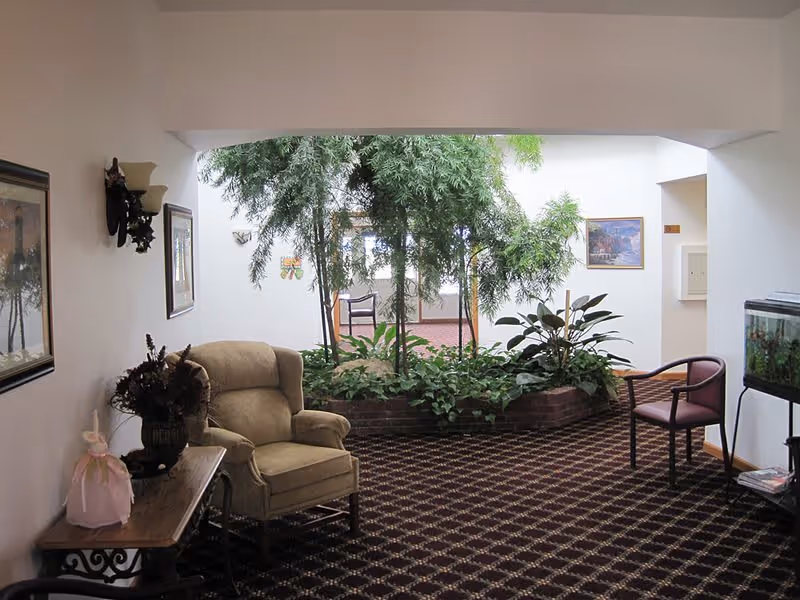 Indoor seating area with a beige armchair, a wooden side table with a decorative item, a patterned carpet, and a large planter with green plants under a skylight. There is also a fish tank on a stand and framed pictures on the walls.