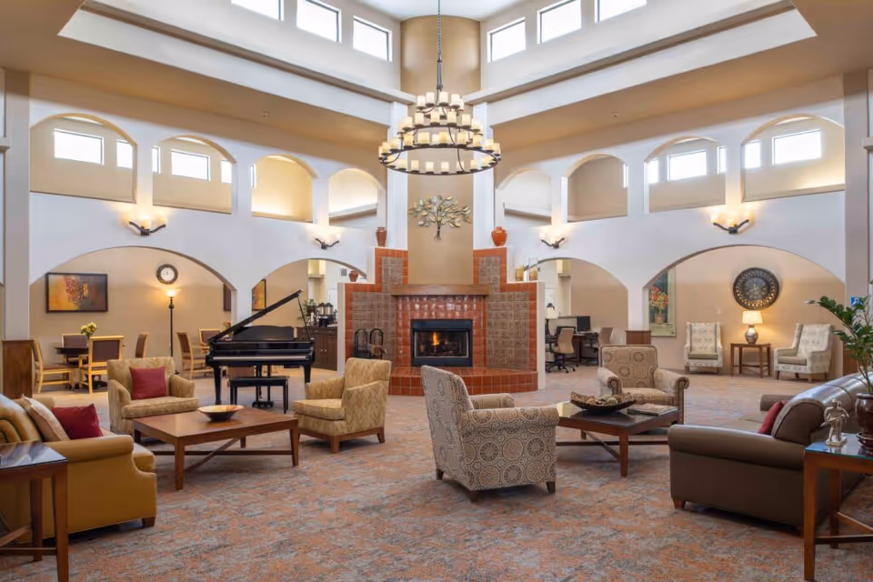 Spacious senior living facility common area with high ceilings and multiple windows. The room features a central fireplace with decorative tiles, surrounded by comfortable armchairs and sofas arranged around coffee tables. A grand piano is positioned near the fireplace. The walls have arches leading to other rooms, and there are various lamps, plants, and wall decorations enhancing the cozy atmosphere.