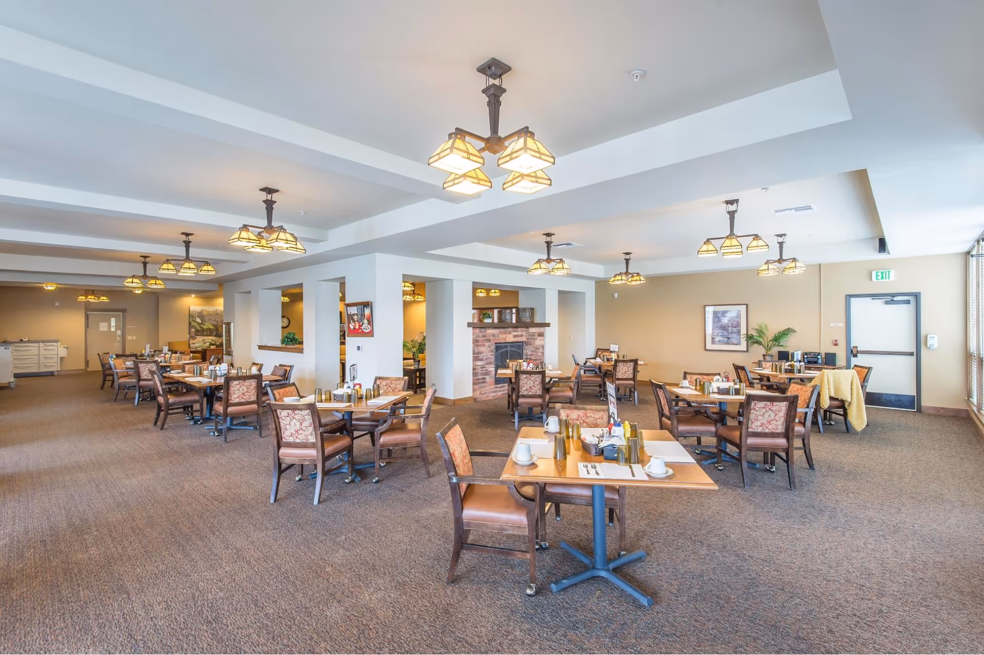 A spacious dining room in an assisted living facility with multiple wooden tables and cushioned chairs arranged neatly. The tables are set with cups, utensils, and condiments. The room features beige walls, carpeted floors, multiple ceiling light fixtures, a brick fireplace, and large windows allowing natural light to enter.