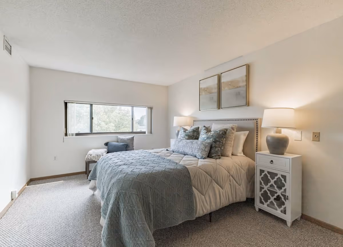 A bright bedroom with a large bed covered in a beige comforter and blue quilt, multiple decorative pillows, two framed artworks above the headboard, a white nightstand with a lamp, and a window with a view of trees.