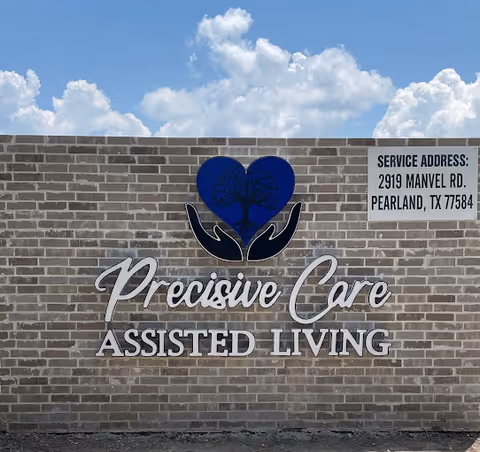 A brick wall with a sign for Precise Care Assisted Living featuring a blue heart with a tree inside held by two black hands. To the right of the sign, there is a service address: 2919 Manvel Rd., Pearland, TX 77584. The sky is partly cloudy in the background.