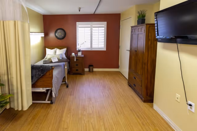 A senior living facility bedroom with a single bed against a red accent wall, a bedside table with a phone and a plant, a wooden wardrobe, a wall-mounted TV, and a window with white shutters. The room has wood flooring and a privacy curtain partially visible on the left side.