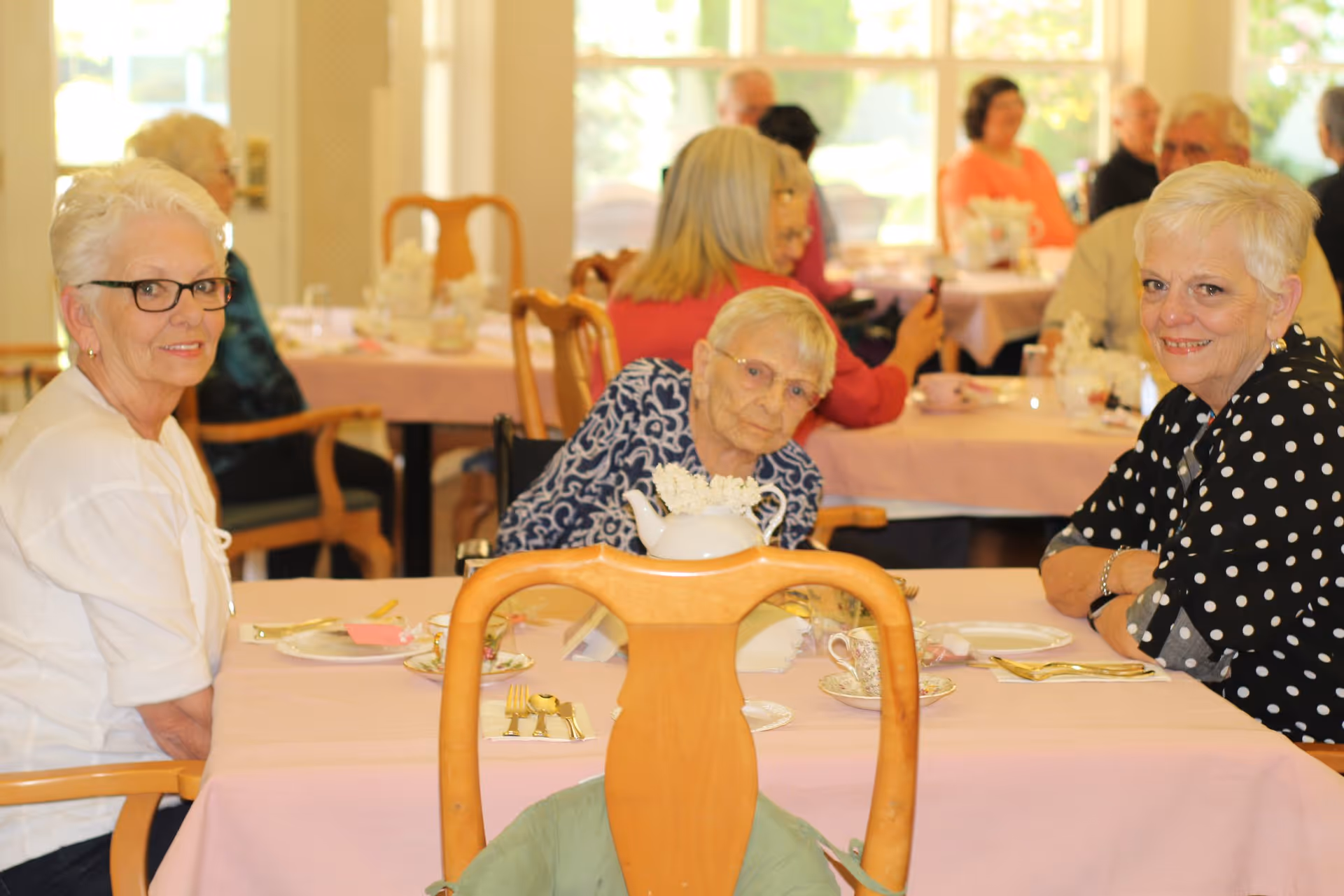 Several elderly women sitting at tables covered with pink tablecloths in a dining room, engaging in conversation and enjoying a meal together. The room is well-lit with large windows in the background.