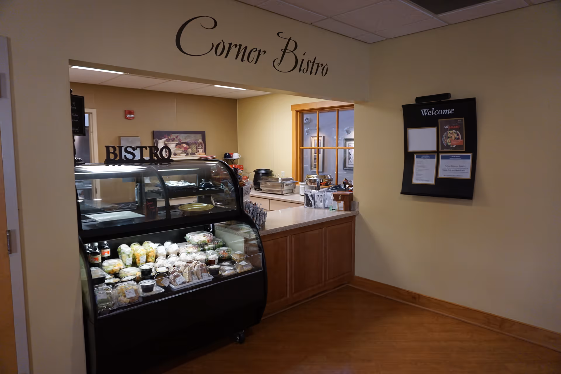 Interior view of a small food service area labeled 'Corner Bistro' with a glass display case containing various packaged food items. There is a counter behind the display case with additional food service equipment and a bulletin board on the wall with a welcome sign and posted notices.
