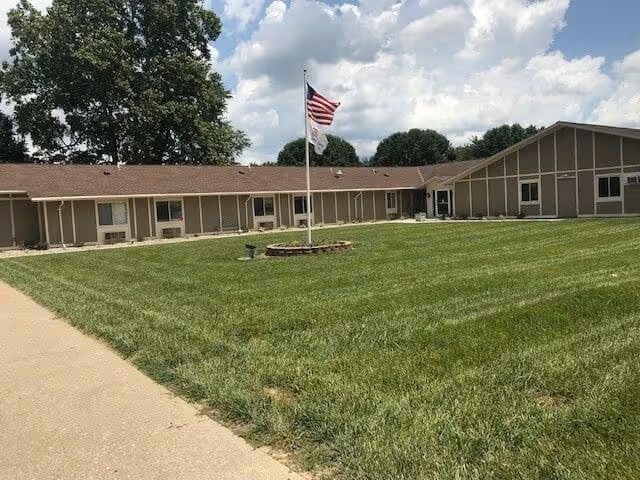 Single-story building with a brown roof and beige walls surrounding a large grassy courtyard with an American flag on a flagpole in the center. Trees and a partly cloudy sky are visible in the background.