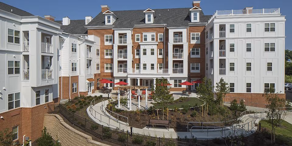 Exterior view of a multi-story brick-and-white senior living building with balconies surrounding a landscaped courtyard with walkways, benches and red patio umbrellas.