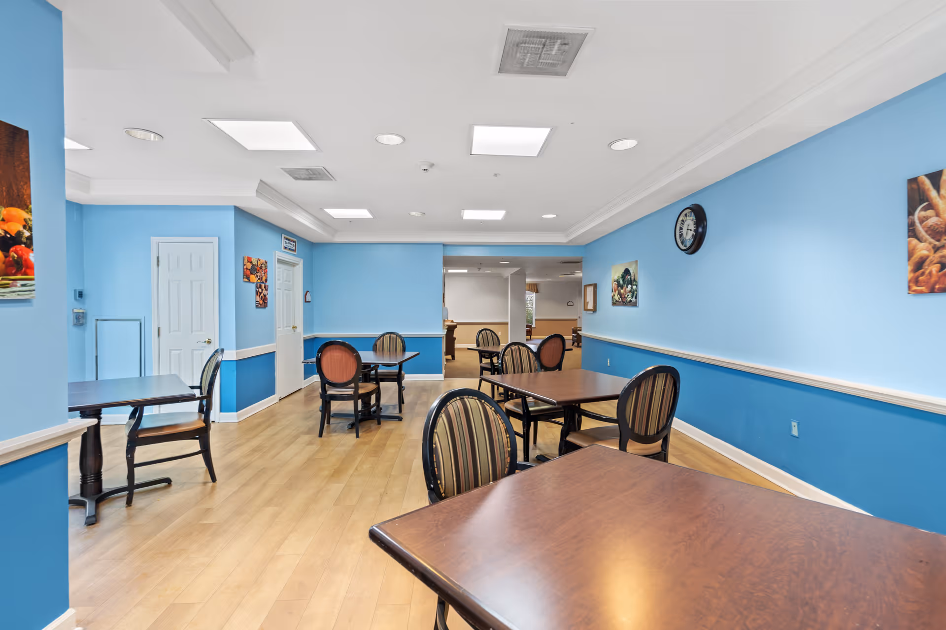 A dining room with light blue walls and wooden flooring. Several tables with chairs are arranged throughout the room. The ceiling has recessed lighting and a clock is mounted on one wall. There are framed pictures of food on the walls.