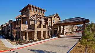 Stone-faced multi-story senior living building with a covered porte-cochere entrance and driveway under a clear blue sky.