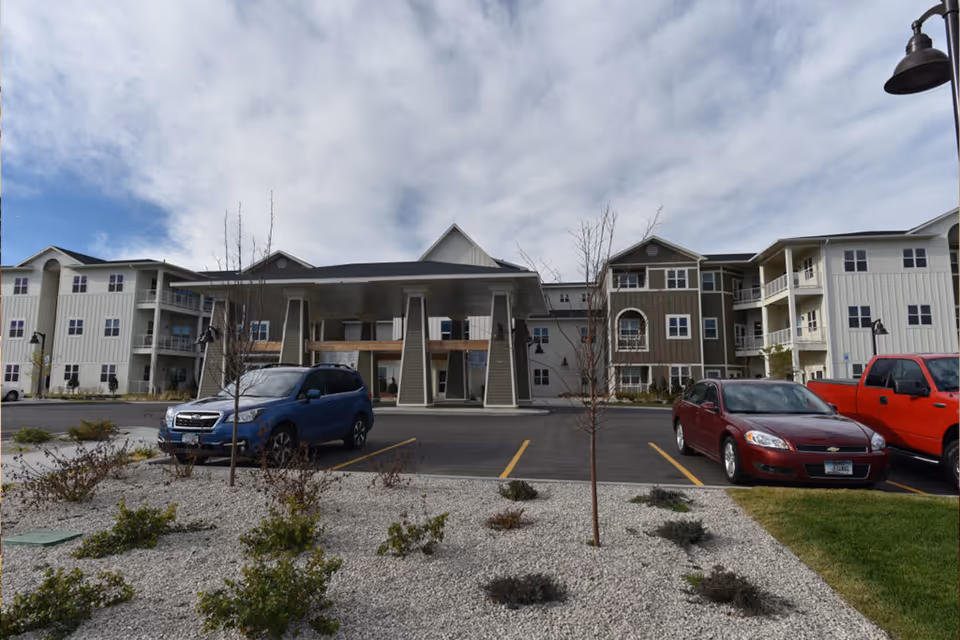 Exterior view of a senior living facility named WyndStone with a covered entrance, multiple floors, balconies, and a parking lot with several cars parked. The sky is partly cloudy and there are small landscaped areas with shrubs and young trees in front.