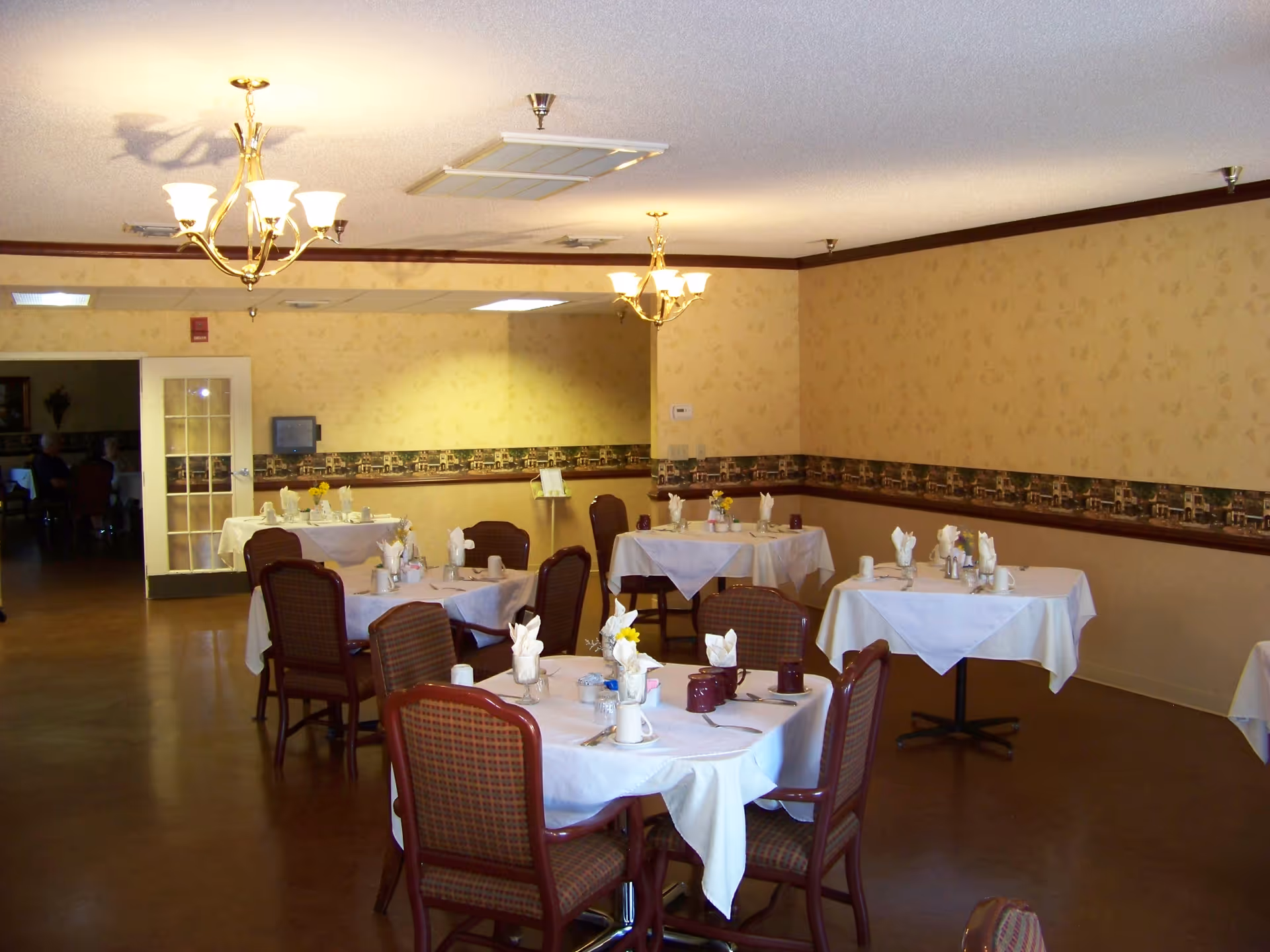A dining room with several round tables covered with white tablecloths, each set with cups, napkins, and utensils. The room has beige wallpaper with a decorative border, wooden chairs with patterned upholstery, and two chandeliers hanging from the ceiling. A large mirror covers one wall, reflecting the room.