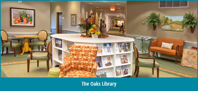 A cozy library-style common area with upholstered chairs, small tables, a magazine-filled shelving unit, and a hallway in the background.