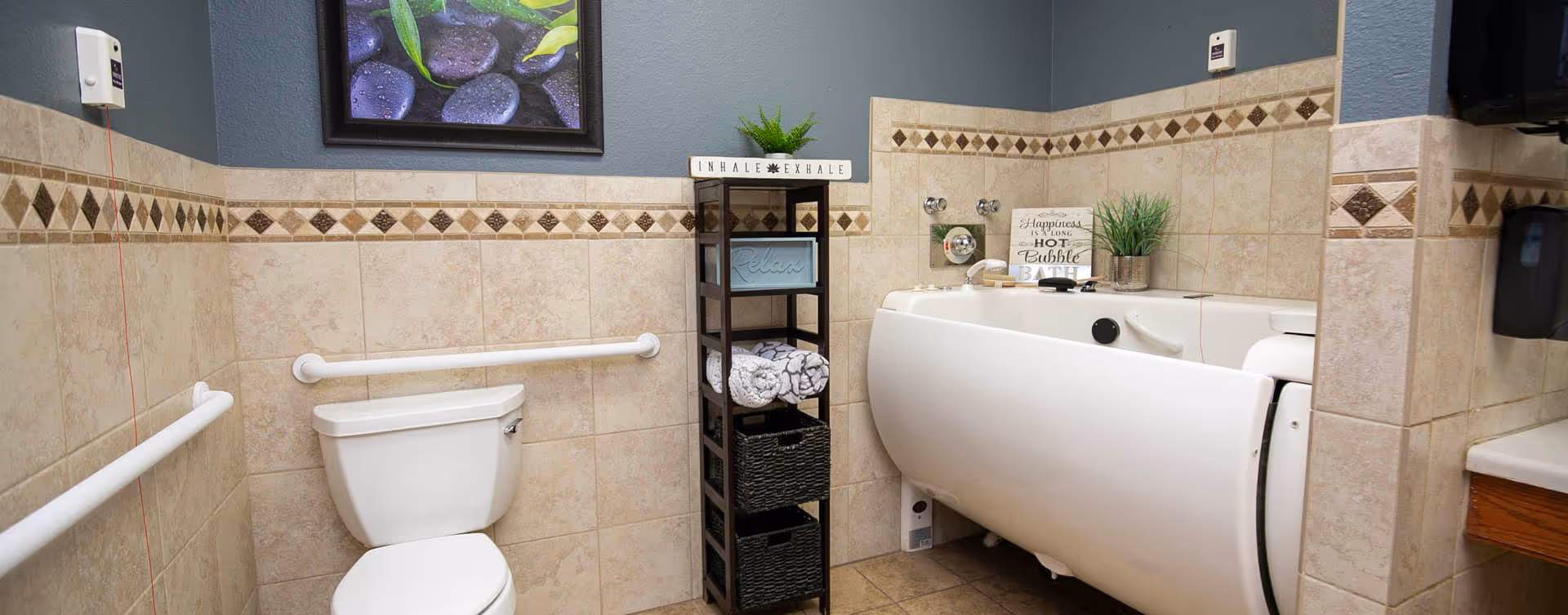 A bathroom with beige tiled walls and floor, featuring a white toilet with grab bars on both sides, a white walk-in bathtub with faucet and controls, and a black shelving unit holding rolled towels and decorative items. A framed picture of stones and leaves hangs on the wall above the toilet.