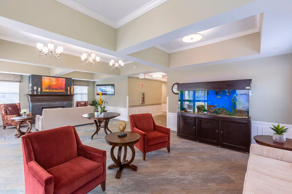 A spacious senior living facility common area with red armchairs, wooden tables, a large aquarium, a fireplace with a colorful painting above it, and a wall-mounted TV. The room has soft lighting from chandeliers and ceiling fixtures, with neutral-colored walls and carpeted floors.
