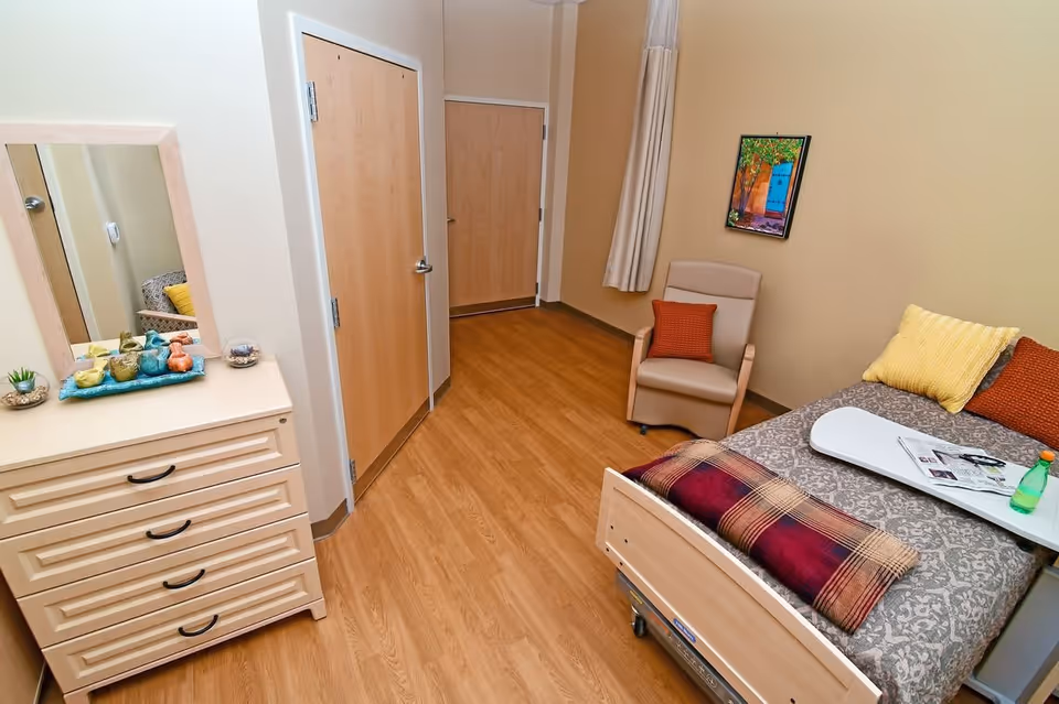 A small, cozy bedroom in a senior living facility featuring a single bed with patterned bedding, a plaid blanket, and colorful pillows. Next to the bed is a beige armchair with an orange cushion. A wooden dresser with a mirror on top holds decorative items. The room has wooden flooring, beige walls, and two closed wooden doors.
