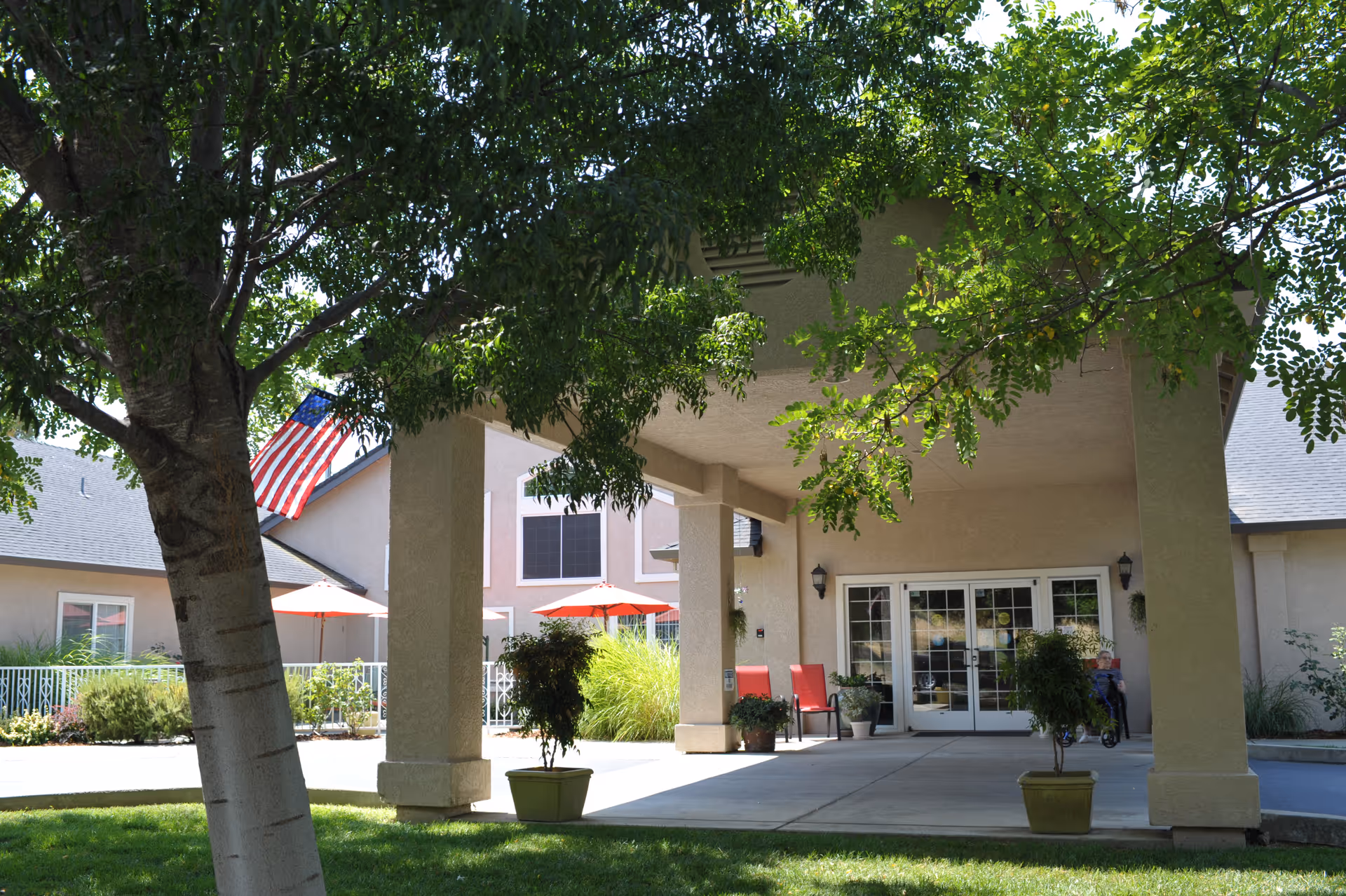 Covered entrance of a senior living building with columns, potted plants, an American flag, and patio seating under red umbrellas.