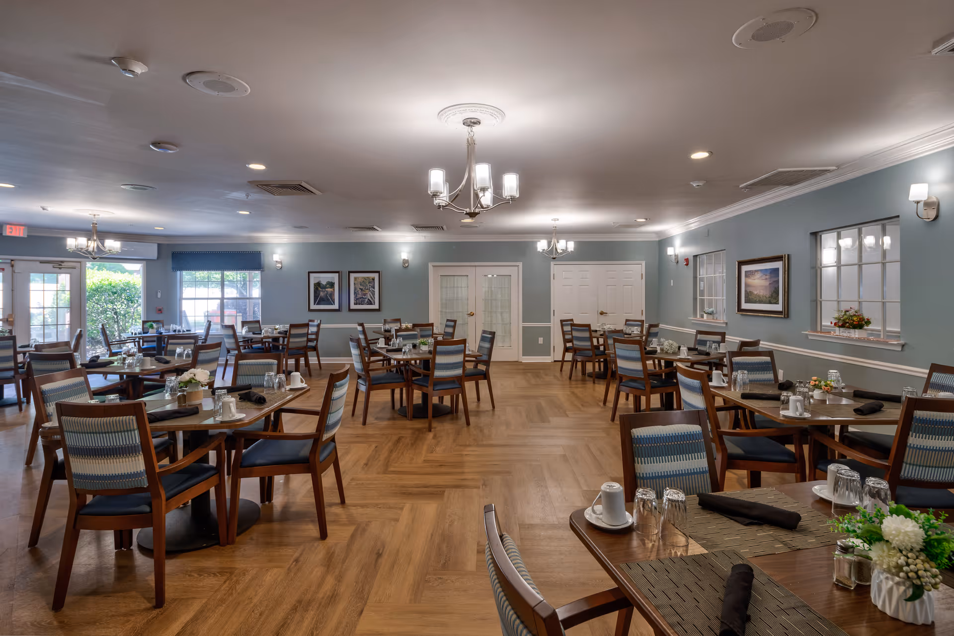 A spacious dining room with multiple wooden tables and chairs arranged neatly. Each table is set with glasses, cups, and black napkins. The room has light blue walls adorned with framed pictures and windows with white trim. There are chandeliers and wall sconces providing warm lighting, and the floor has a wood pattern. A door and windows are visible at the far end, letting in natural light.