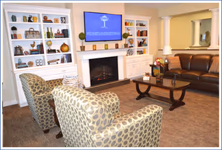 A cozy living room with two patterned armchairs and a brown leather sofa arranged around a wooden coffee table. Behind the seating area is a white fireplace with a flat-screen TV mounted above it, flanked by built-in white bookshelves filled with decorative items. A large mirror hangs on the wall above the sofa.