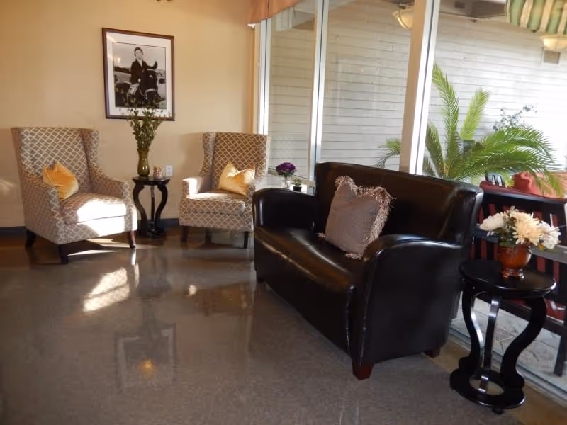 A cozy seating area with two patterned armchairs with yellow cushions, a black leather loveseat with a decorative pillow, and two small black side tables with flower arrangements. A large window behind the loveseat shows some green plants outside. A framed black and white photograph hangs on the beige wall.