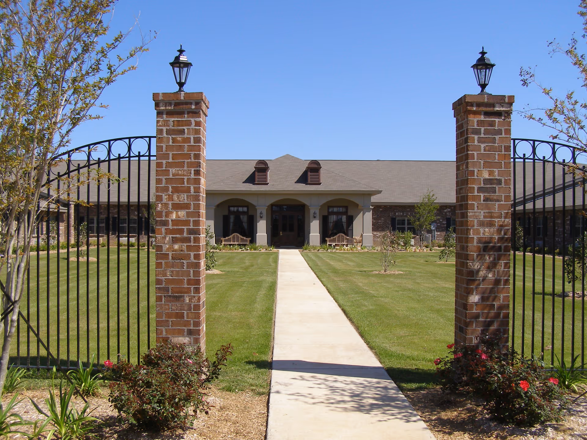 View of the front exterior of Senior Village Nursing & Rehab Center showing a paved walkway leading through a black iron gate with brick pillars and lanterns to the entrance of a single-story building with a pitched roof, surrounded by a well-maintained lawn and some small trees and bushes.