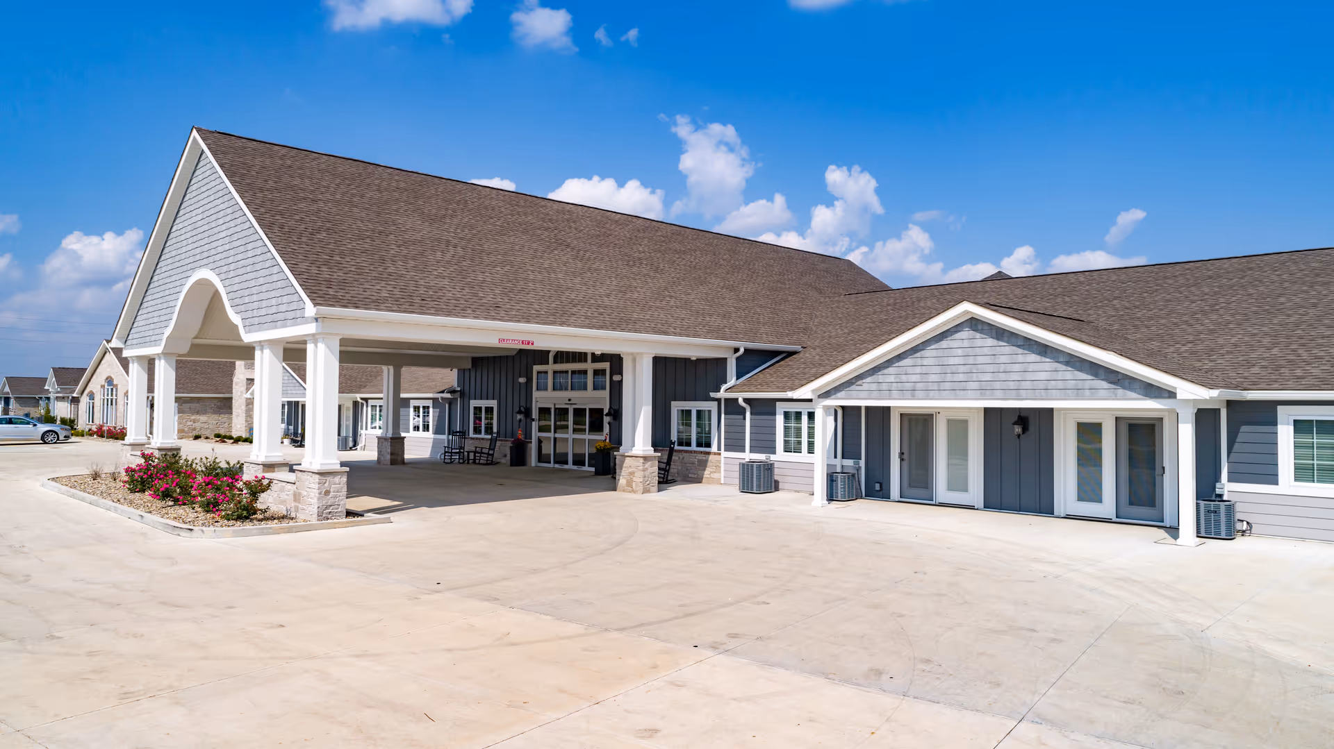 Exterior view of a single-story assisted living facility building with a large covered entrance, gray siding, white trim, and a brown shingled roof under a blue sky with scattered clouds. There are some flower beds with pink flowers near the entrance and a few parked cars visible in the background.