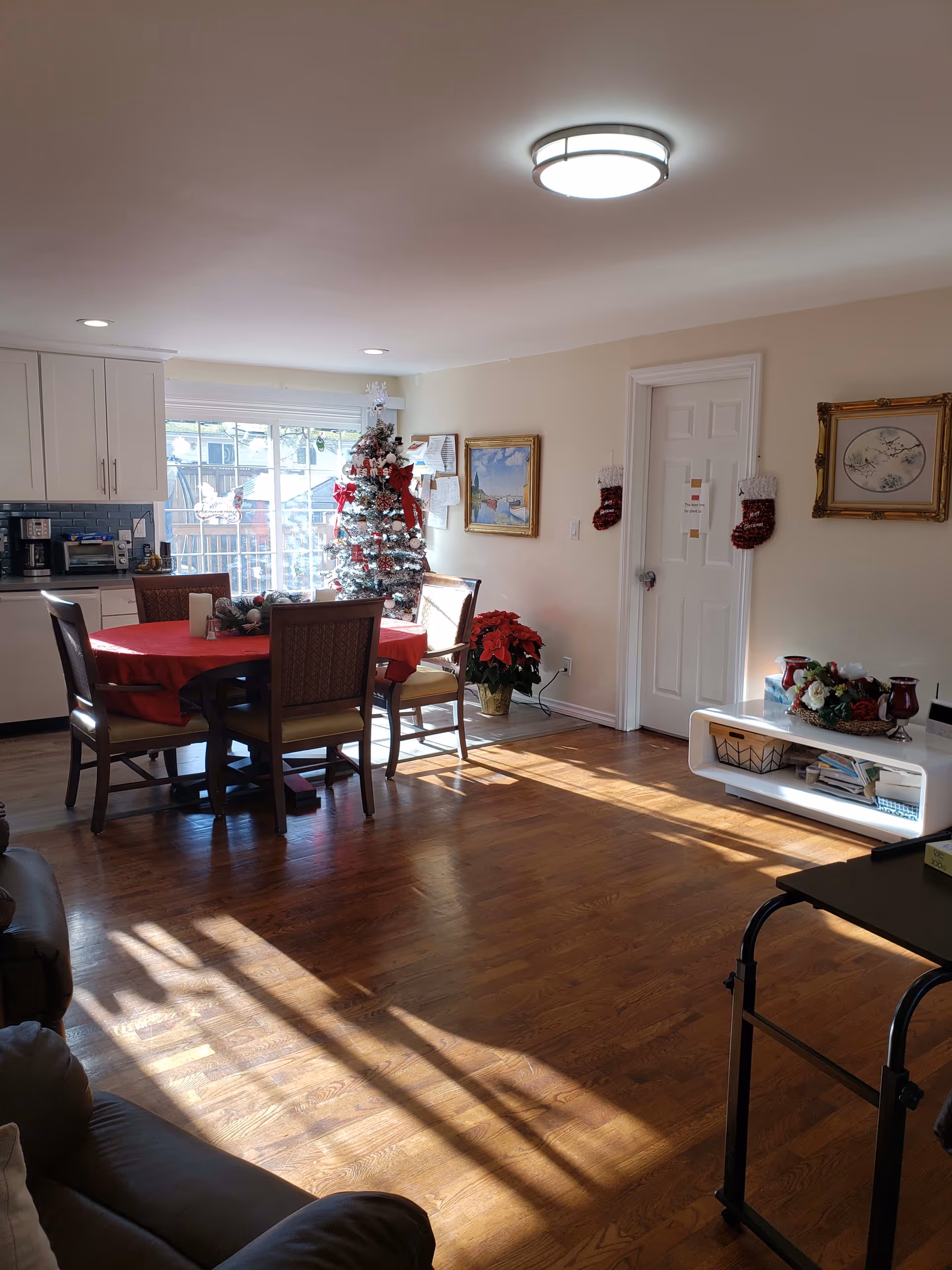 A bright living and dining area with wooden floors, a round dining table covered with a red tablecloth and four chairs, a decorated Christmas tree near a large window, a white cabinet with kitchen appliances, and a white door with Christmas stockings hanging on either side. There are framed paintings on the walls and a white shelf with decorative items and books.