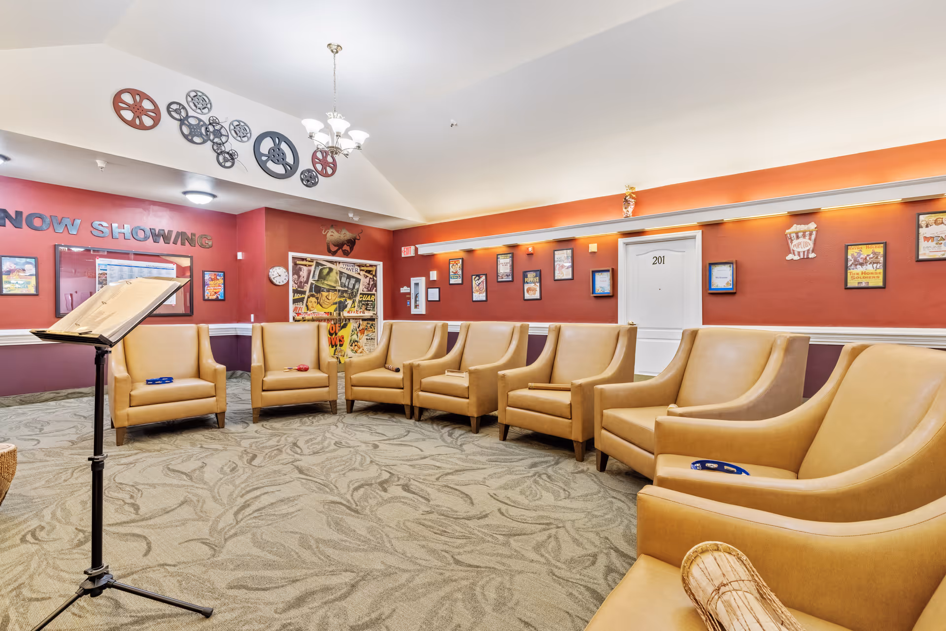 A cozy common area with a semi-circle arrangement of tan leather armchairs on a patterned carpet. The walls are painted red and decorated with framed movie posters and film reel wall art. A music stand is positioned in the foreground, and a door labeled '201' is visible on the back wall. The room has a warm, inviting atmosphere with soft lighting.