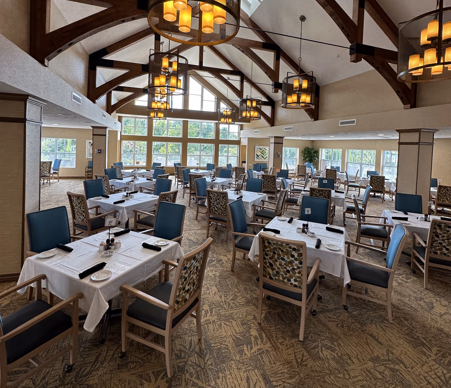 Spacious dining room with multiple set tables and chairs, exposed wooden ceiling beams, large windows, and hanging chandeliers.