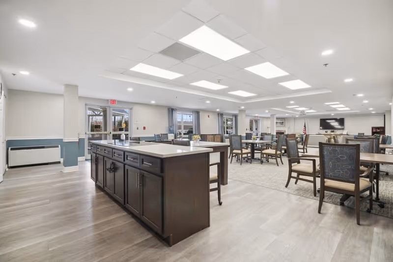 A spacious and well-lit common dining area in a senior living facility featuring multiple tables and chairs arranged for group seating. The room has a large central island with dark cabinetry and a light countertop, wood flooring, and large windows allowing natural light. The ceiling has recessed lighting panels, and there is a television mounted on the far wall.
