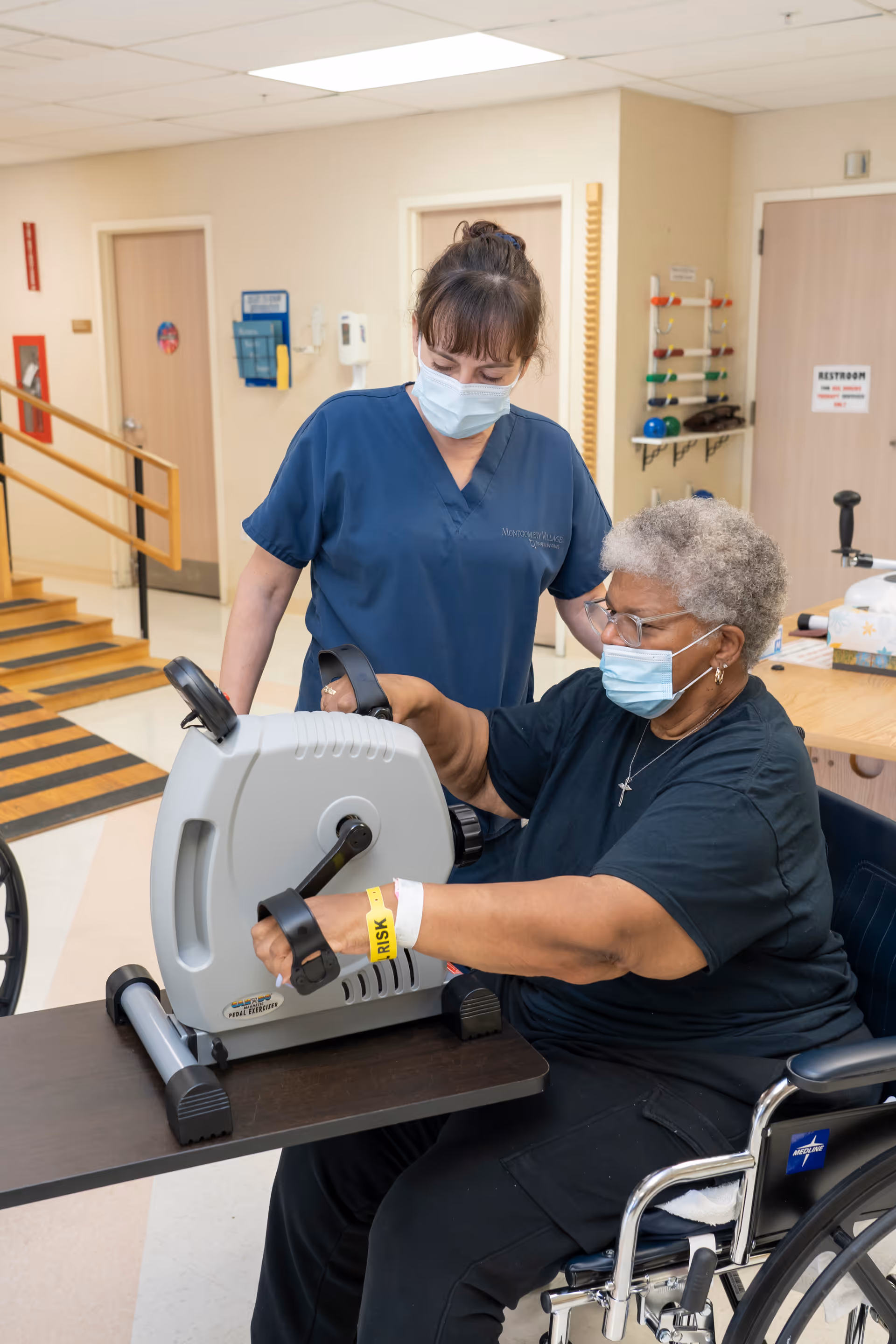 A healthcare worker wearing blue scrubs and a face mask assists an elderly woman in a wheelchair who is exercising her arms using a hand pedal exerciser in a healthcare facility hallway.