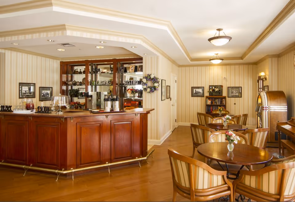 A cozy dining area with round wooden tables and striped upholstered chairs. A wooden counter with coffee dispensers and a glass cake stand is on the left side. The walls are decorated with framed pictures and a floral wreath, and there is a vintage jukebox in the corner. The room has warm lighting and a wooden floor.