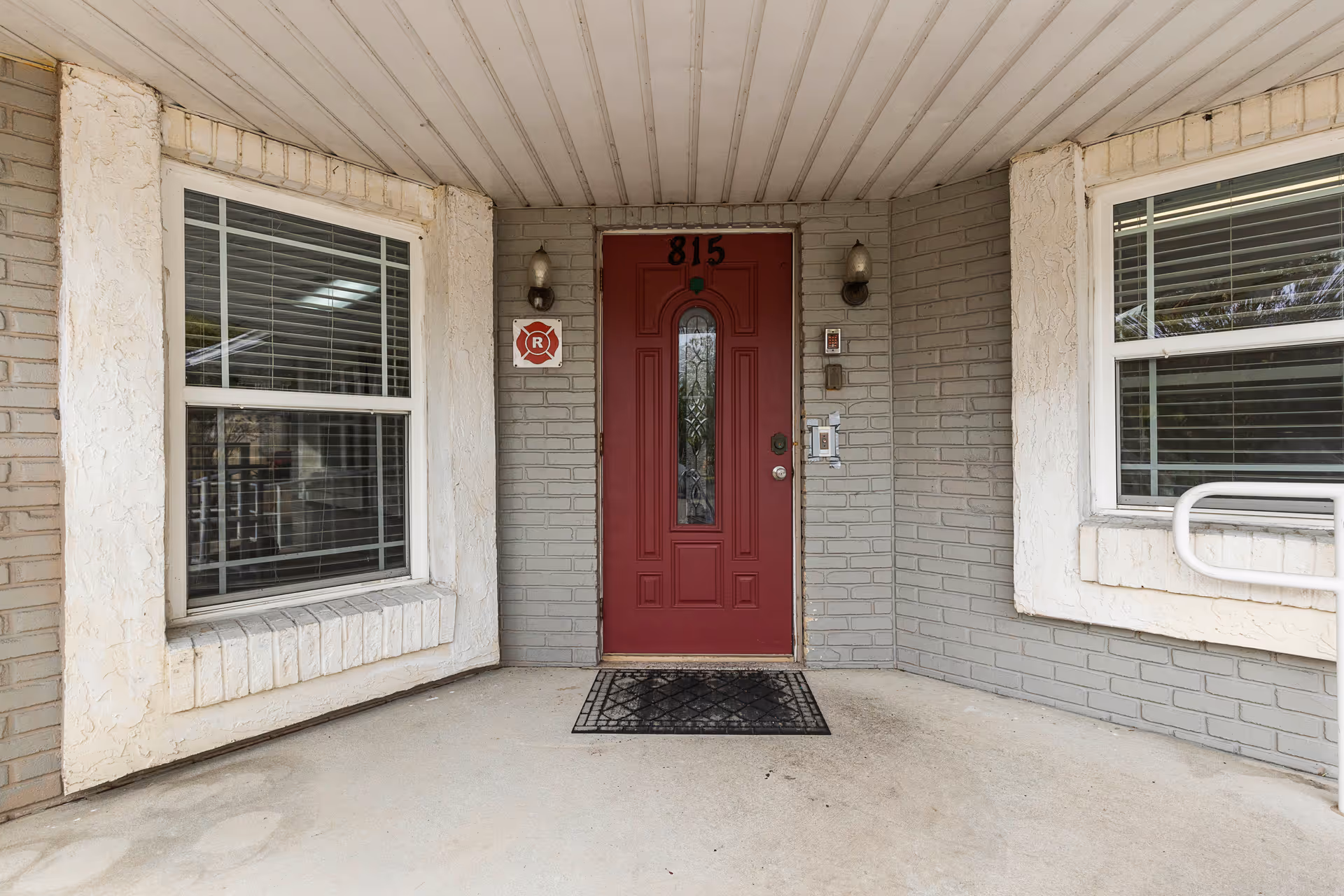 Covered entrance area of a building with a red front door numbered 815, flanked by two windows with white frames and blinds. The walls are painted gray brick with white textured trim around the windows. There are two wall-mounted light fixtures on either side of the door and a black doormat in front of the door.